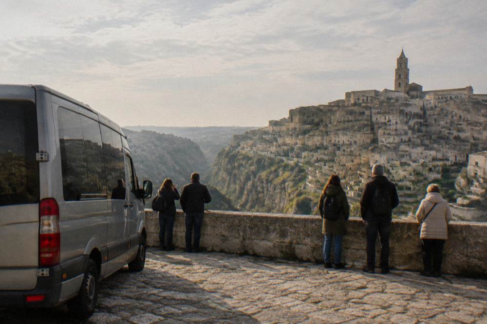 A tour minibus parked at a belvedere facing tiers of honey-coloured cave dwellings across a ravine, visitors seen from behind at a low wall, hazy morning, Matera