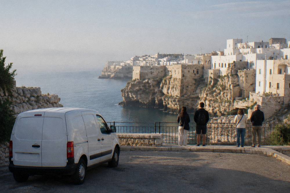 A small white van parked at a cliff-edge viewpoint, a small group seen from behind at the railing looking at a white town on limestone cliffs, bright light, Polignano a Mare