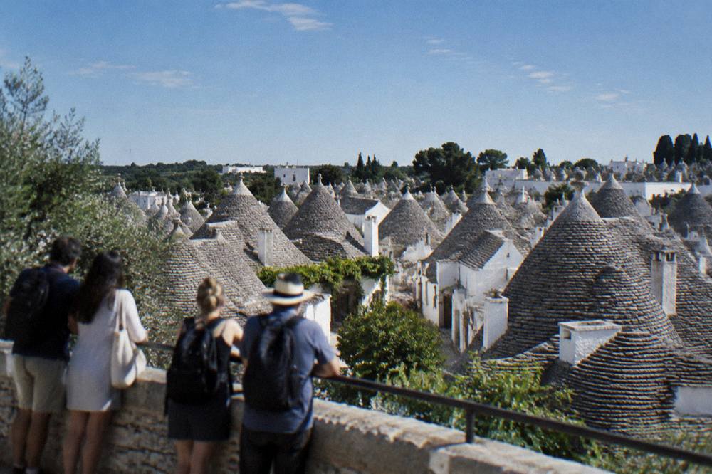 A wide view over a dense cluster of conical gray stone roofs from a belvedere, a small group at the railing seen from behind, Alberobello
