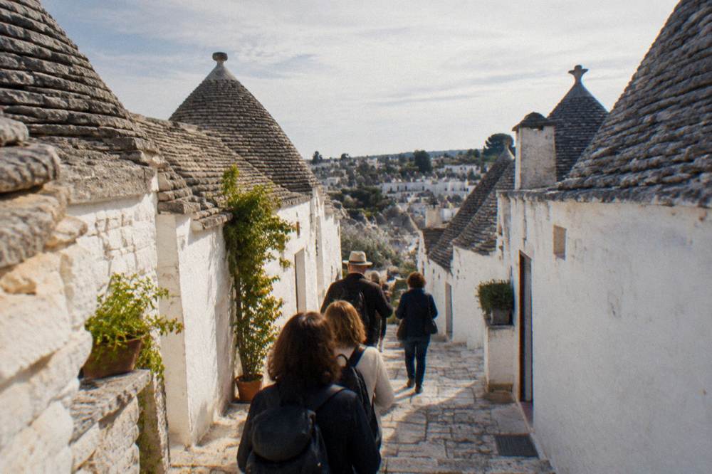 A small group seen from behind walking down a stepped lane between white houses with gray conical roofs, a guide ahead, bright light, Alberobello