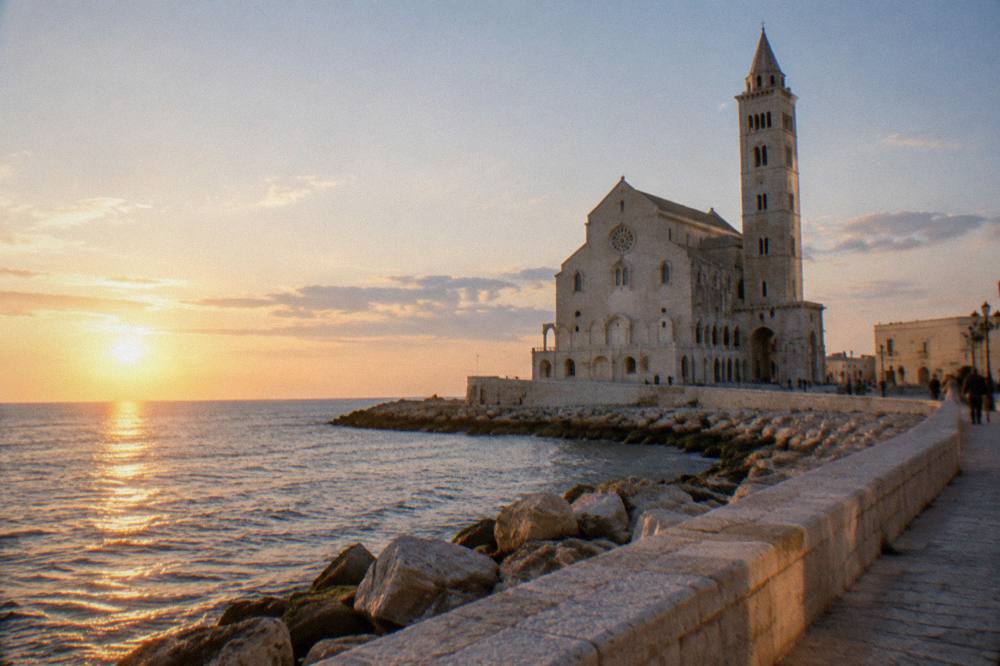 A Romanesque cathedral of pale limestone on the edge of the sea with a tall slender bell tower, visitors far off, bright light, Trani