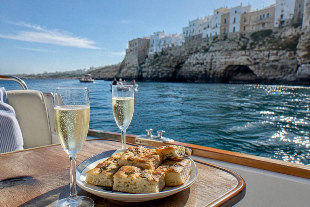 A boat table on deck with two glasses of prosecco and a plate of focaccia, the cliffs of a white town blurred behind, sun on the water, Polignano a Mare