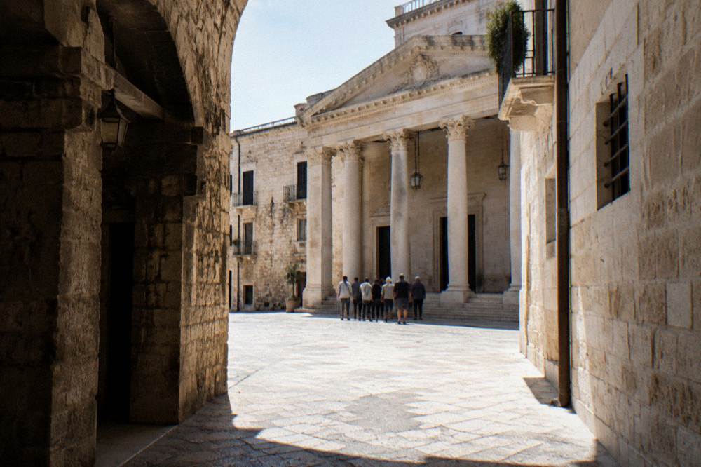 A small group of people far off seen from behind gathering in front of a pale neoclassical theater portico at the start of a walk, morning light, Bari Vecchia