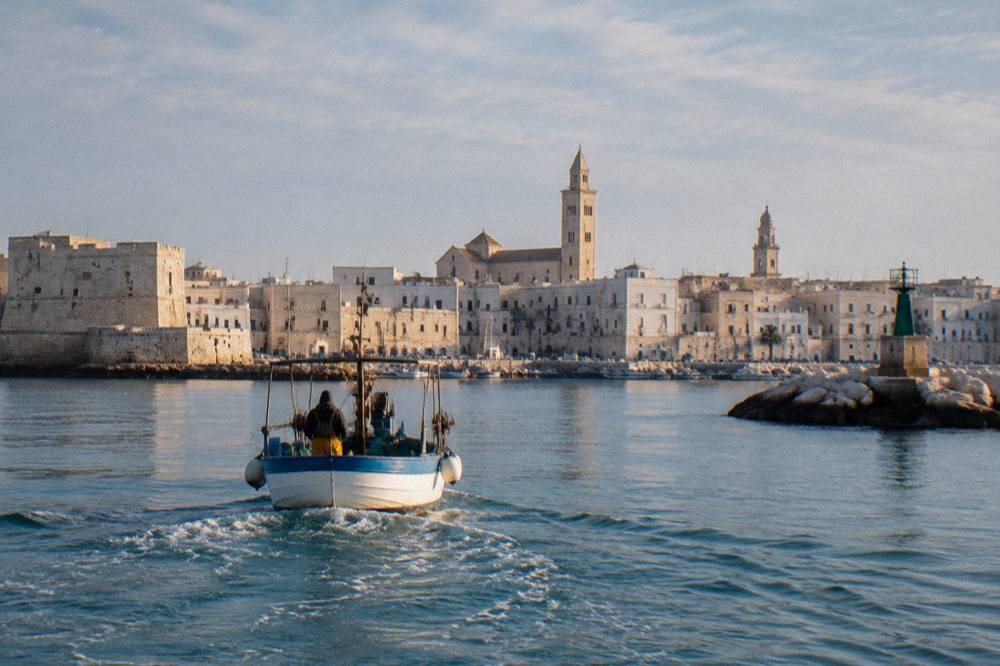 A small boat heading out of the old harbor with the pale old town skyline and two towers behind it, seen from the water, soft morning light, Molo San Nicola