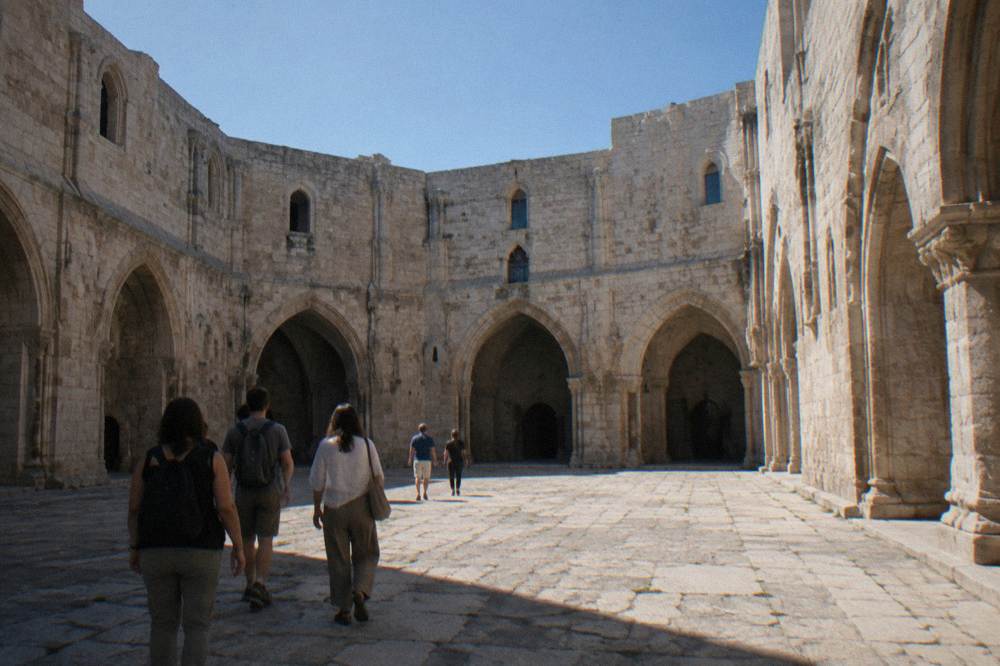 A small group seen from behind in the octagonal courtyard of a medieval castle, arched doorways in pale stone, blue sky overhead, Castel del Monte