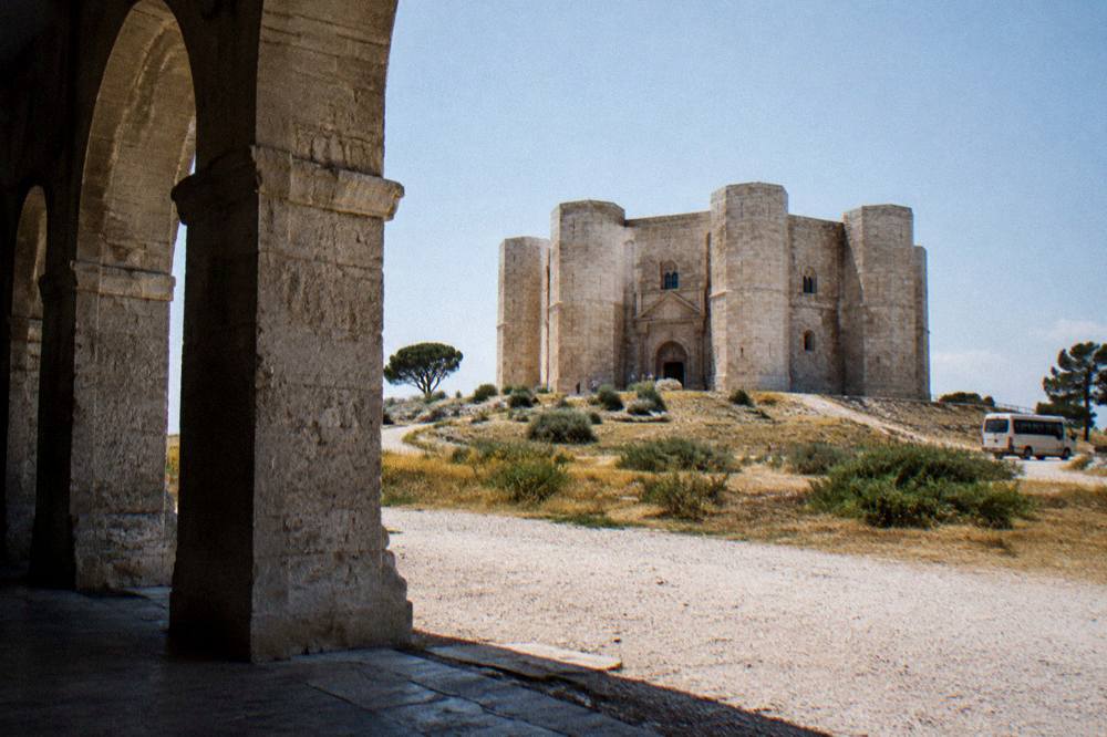 An octagonal pale limestone fortress with eight corner towers on a low hill, a small tour van parked far off on a gravel area, dry fields, clear sky, Castel del Monte