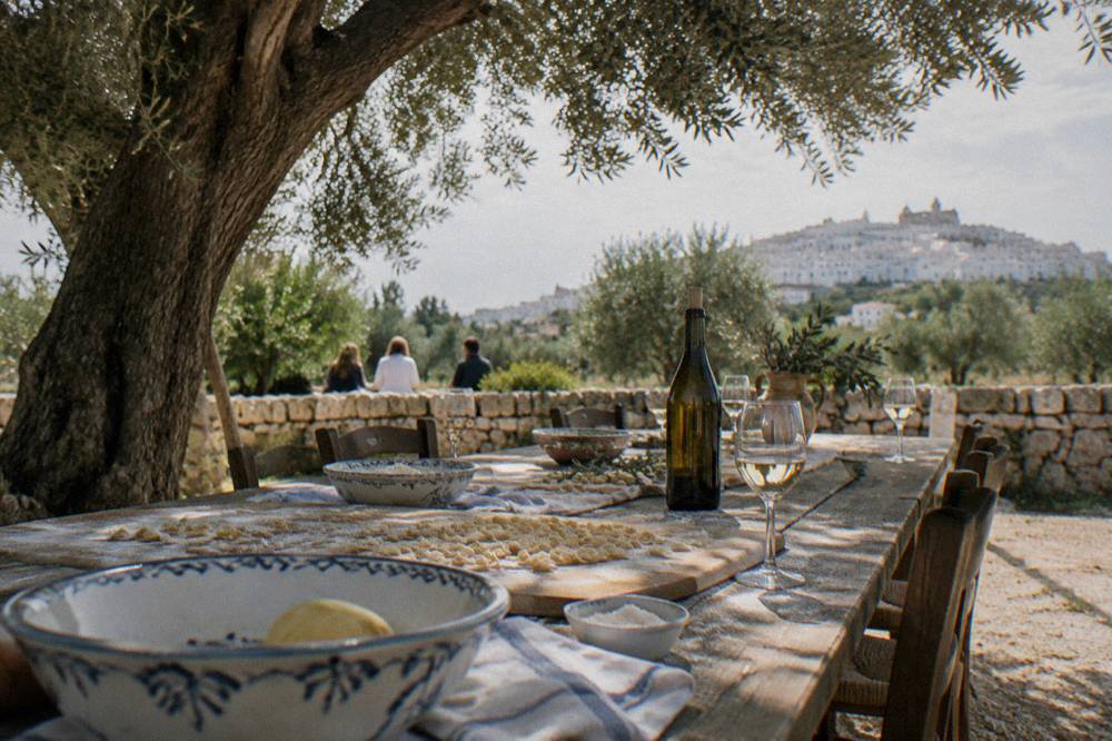 A long outdoor table under an olive tree on a farm with bowls of pasta dough, a wine bottle and glasses, bright dappled light, Ostuni