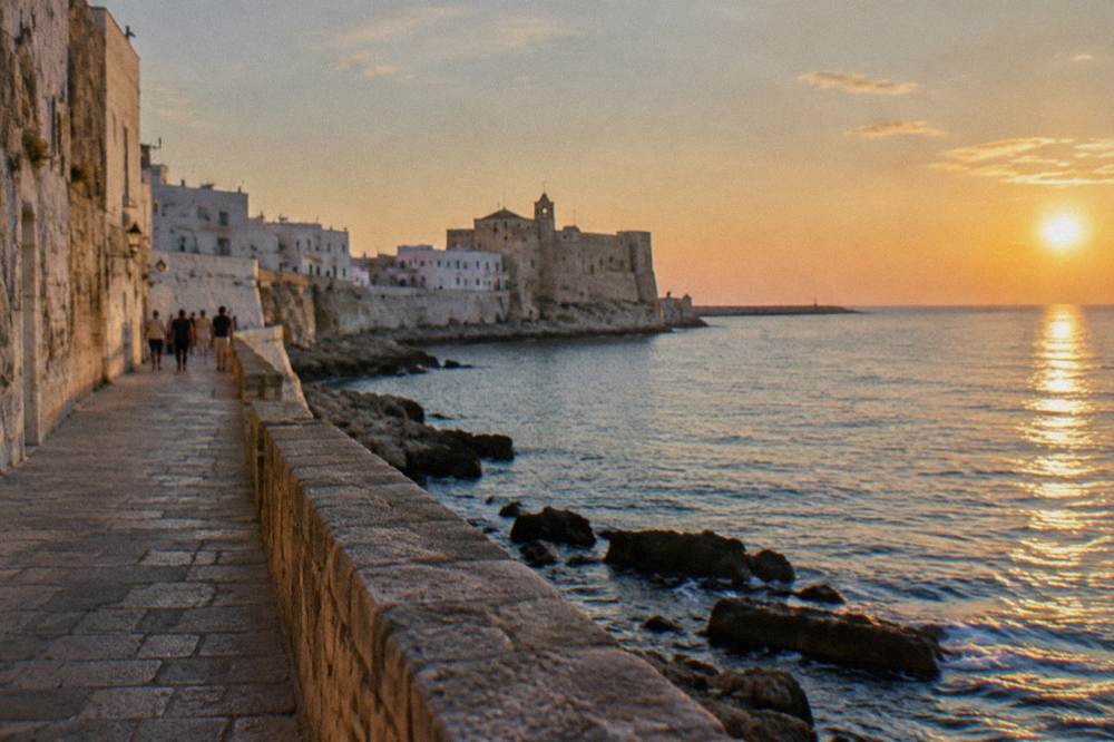 A group far off strolling along a sea promenade beside a whitewashed old town wall, a stone castle on the headland, afternoon, Monopoli