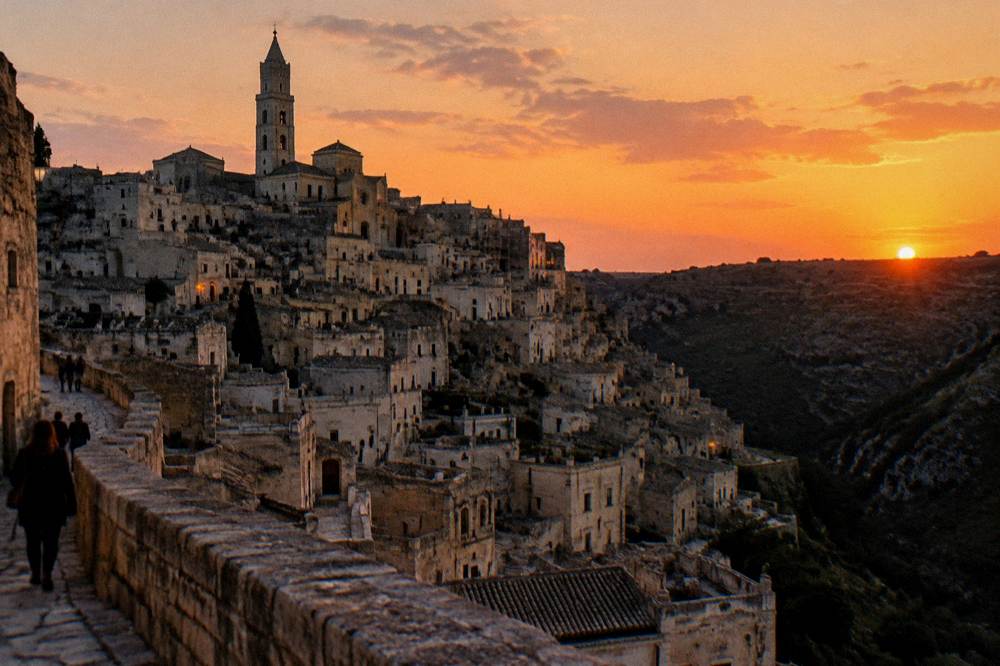 A cathedral bell tower rising over stacked stone houses at sunset, the ravine in shadow, the sky orange, Matera