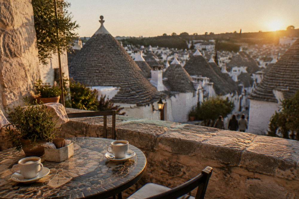 A small stone terrace with a table, two coffee cups and a view of conical roofs below, warm light, Alberobello