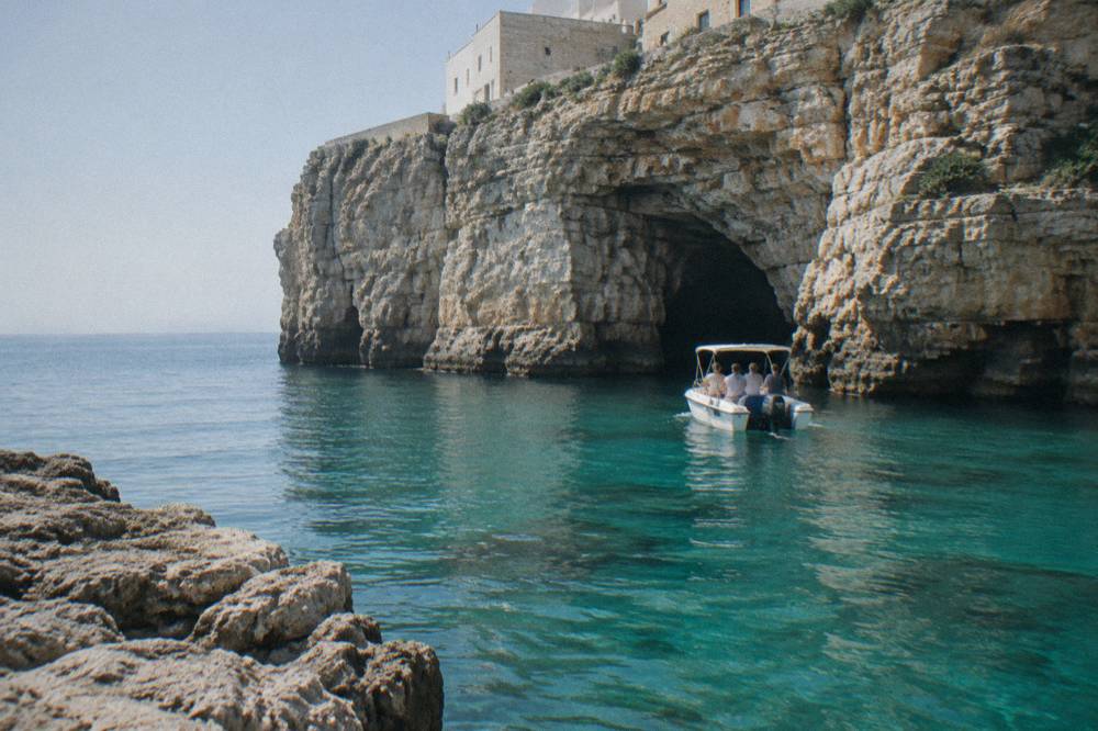 A small motorboat idling near tall pale limestone cliffs and a sea cave opening, turquoise water, a few passengers seen from behind, bright midday, framed wider with more foreground, Polignano a Mare