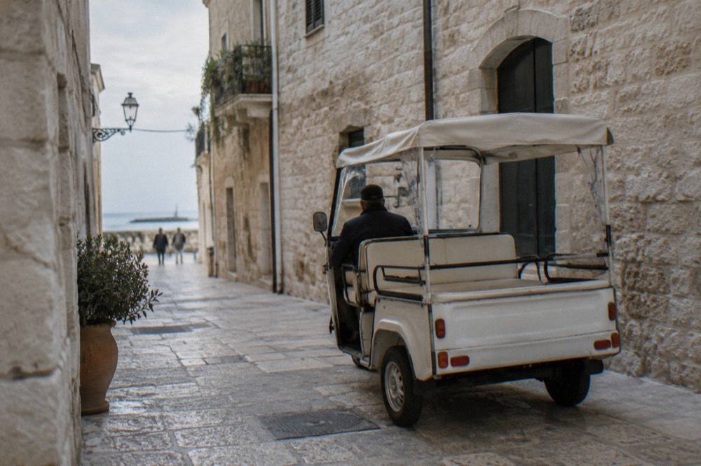 An unmarked three-wheeled electric rickshaw with a canopy parked in an old-town lane of pale limestone, a driver's back turned, soft morning light, Bari Vecchia