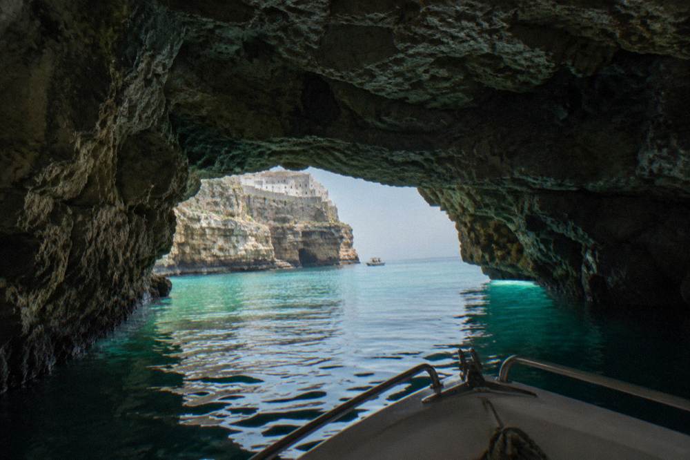 Inside a sea cave looking out through a low arch to bright turquoise water, a boat's bow entering, dim rock walls, Polignano a Mare