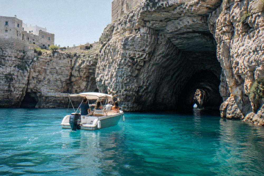 A small motorboat idling near tall pale limestone cliffs and a sea cave opening, turquoise water, a few passengers seen from behind, bright midday, Polignano a Mare