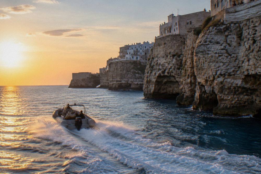 A fast rigid boat leaving a white wake along a wall of pale limestone cliffs, spray catching the sun, the sea deep blue, Polignano a Mare