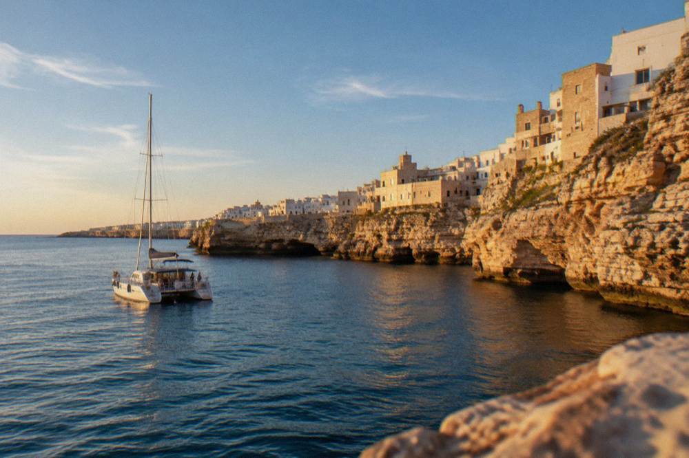 A sailing catamaran with a trampoline net anchored off the cliffs at golden hour, passengers far off, warm light on the rock, Polignano a Mare