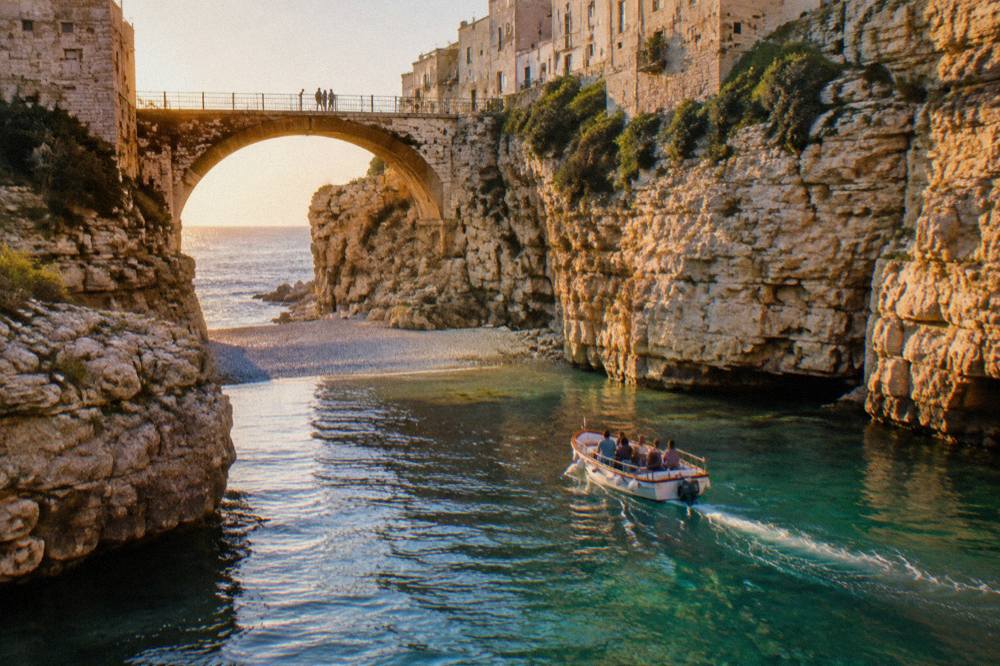 A boat gliding past the mouth of a narrow pebble cove with a low arched bridge high above, clear shallow water, afternoon light, Lama Monachile