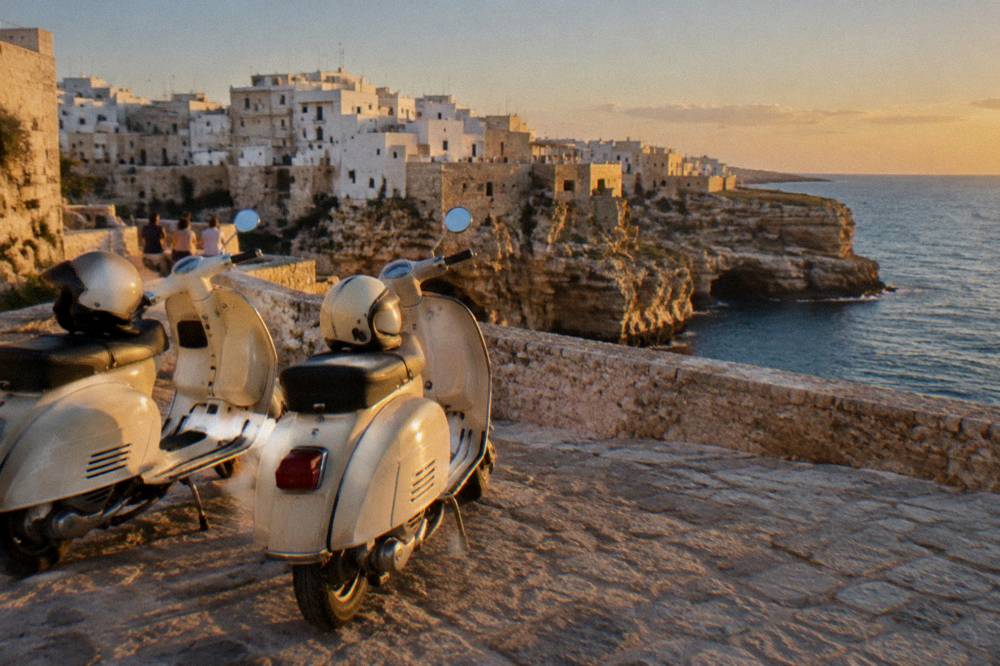 Two cream vintage scooters parked at a cliff-edge viewpoint with a white town on limestone cliffs and blue sea beyond, helmets on the seats, Polignano a Mare