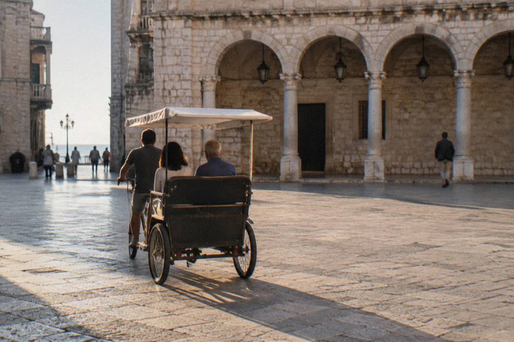 A three-wheeled rickshaw with a plain canopy crossing a pale stone square with a loggia behind, passengers seen from behind, warm light, Piazza Mercantile