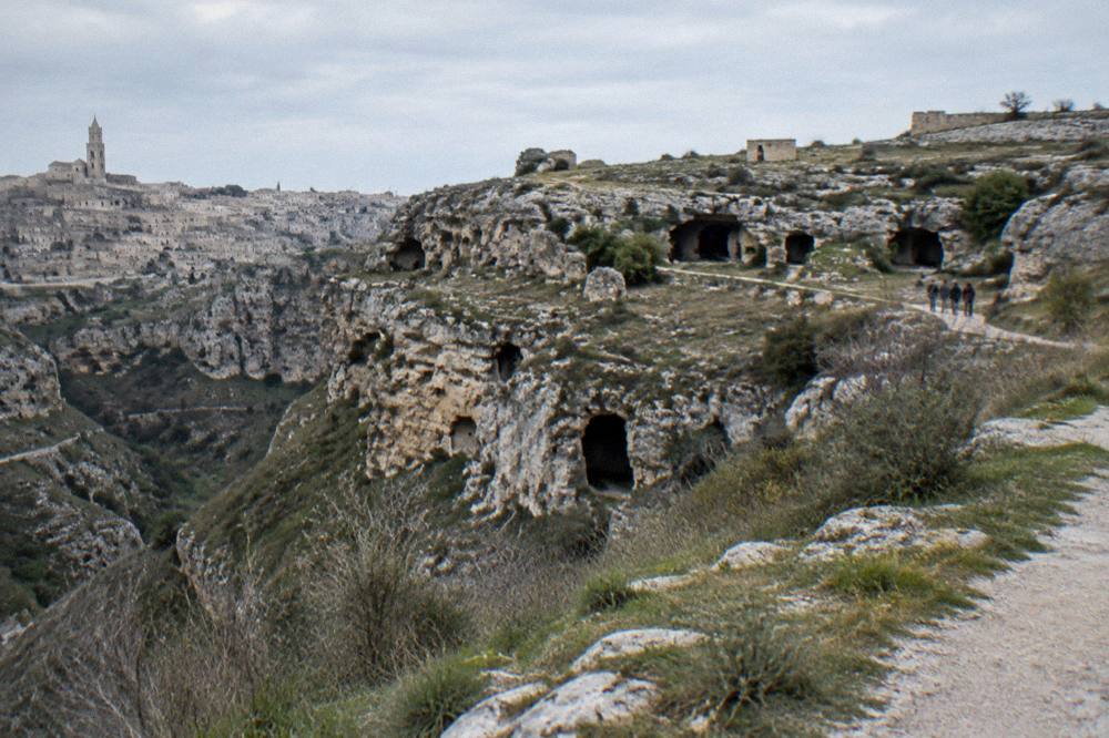 A rocky plateau across a ravine with dark cave openings and sparse scrub, a narrow path, a small group far off, clear late light, Matera
