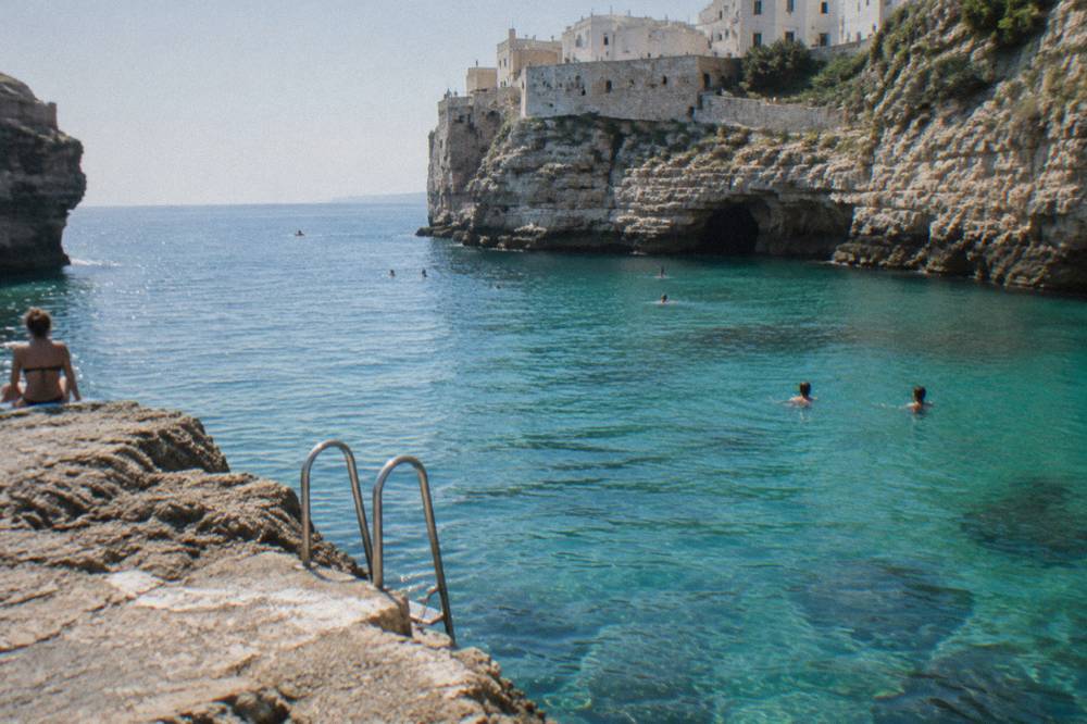 A swimming stop in a turquoise cove under cliffs, a boat ladder into the water, a few distant swimmers, strong sun, Polignano a Mare