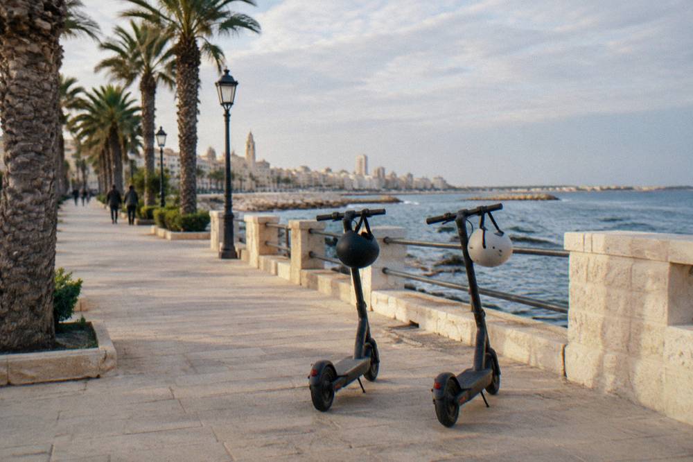 Two upright two-wheeled personal electric scooters parked on a seafront promenade, helmets hung on the handles, palm trees, soft light, Lungomare Nazario Sauro