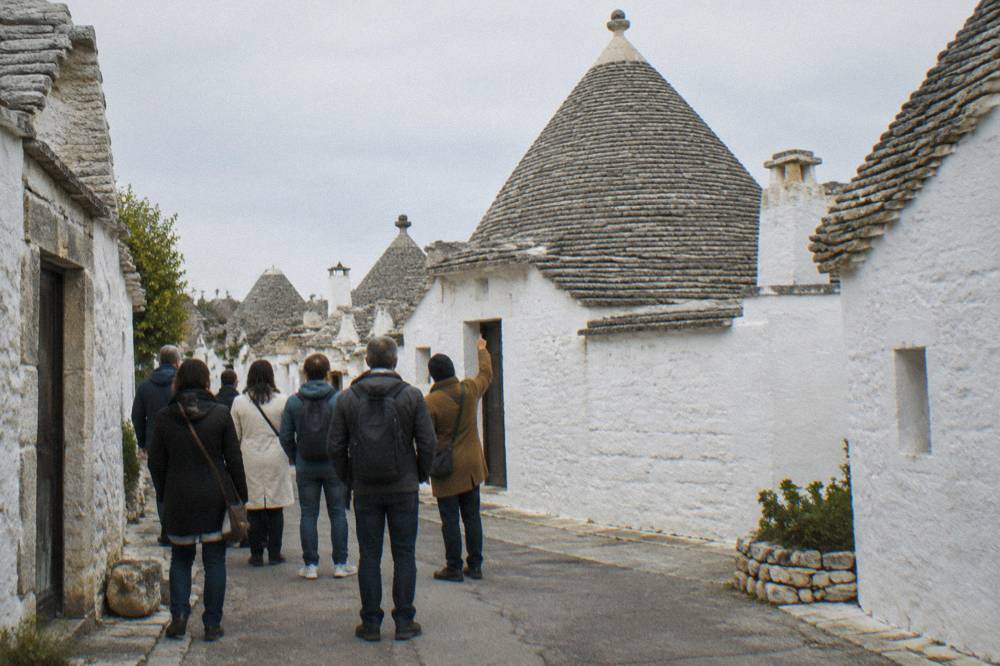 A small group seen from behind in front of a white house with a gray conical roof, a guide pointing, bright light, Alberobello