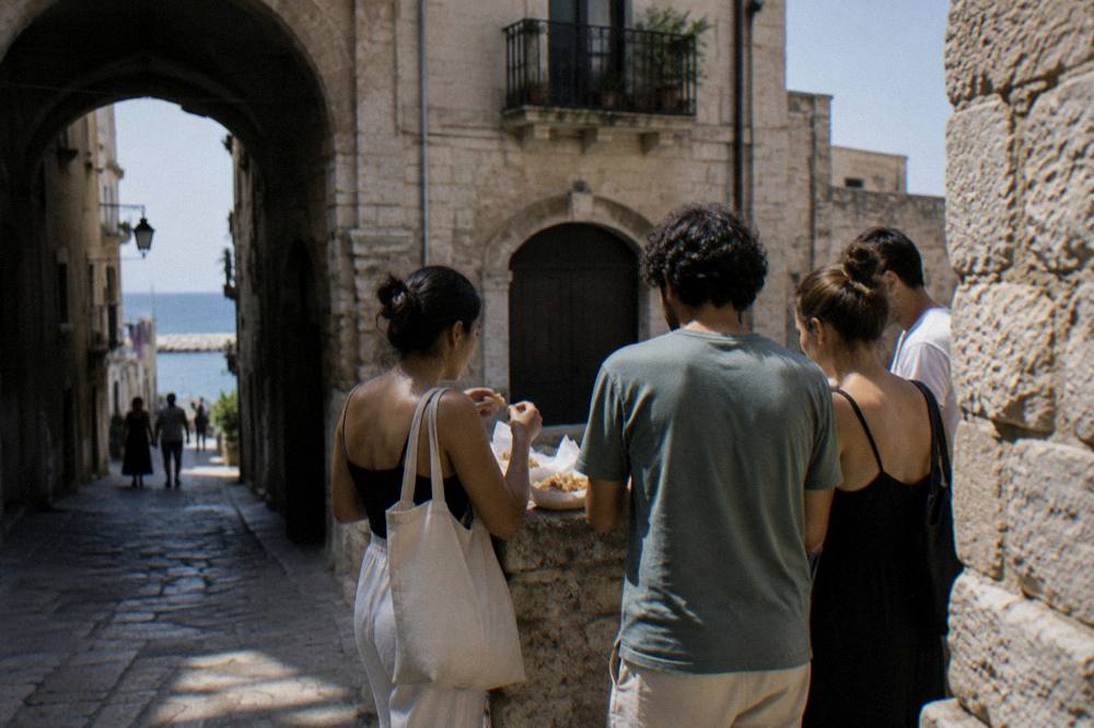 A small group seen from behind gathered round a stone ledge in an old-town lane sharing paper trays of fried street food, soft daylight, Bari Vecchia
