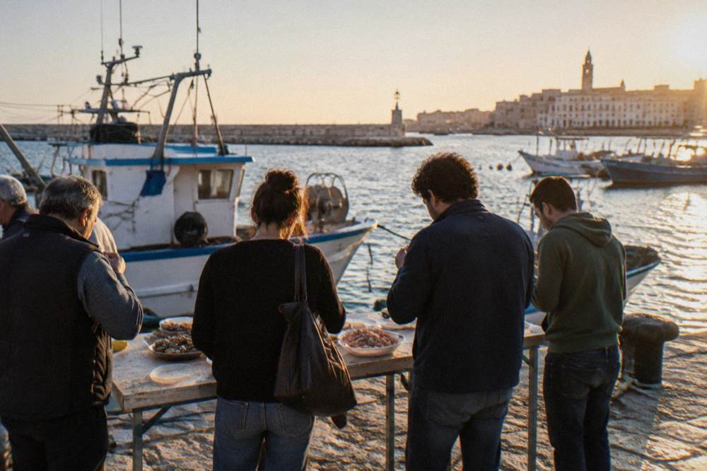 A group seen from behind standing at the edge of a harbor pier tasting raw seafood from a plain table, fishing boats behind, bright sun, Molo San Nicola