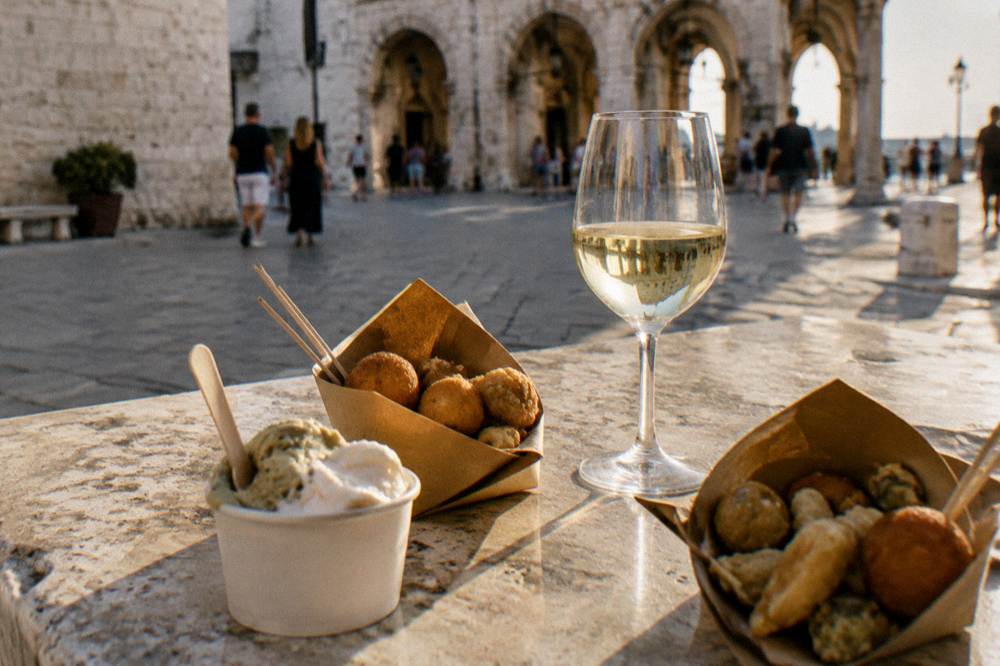 A stone table in a piazza with paper cones of small fried snacks, a gelato cup and a glass of white wine, arches blurred behind, Piazza del Ferrarese