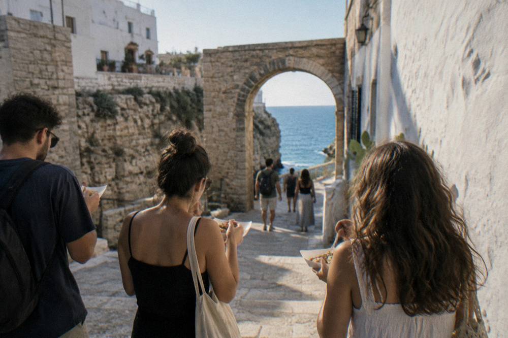 A small group seen from behind eating from paper trays on a whitewashed cliff-top lane, a stone arch and a glimpse of blue sea ahead, Polignano a Mare
