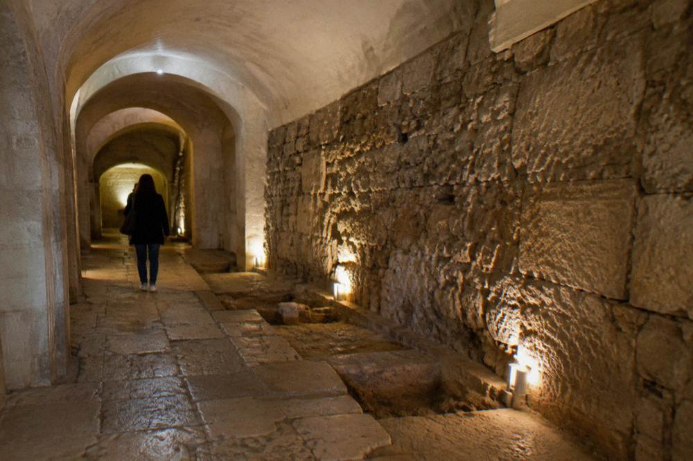 A narrow vaulted underground passage with excavated ancient paving and an exposed old stone wall, soft spot lighting, a visitor seen from behind, Basilica di San Nicola
