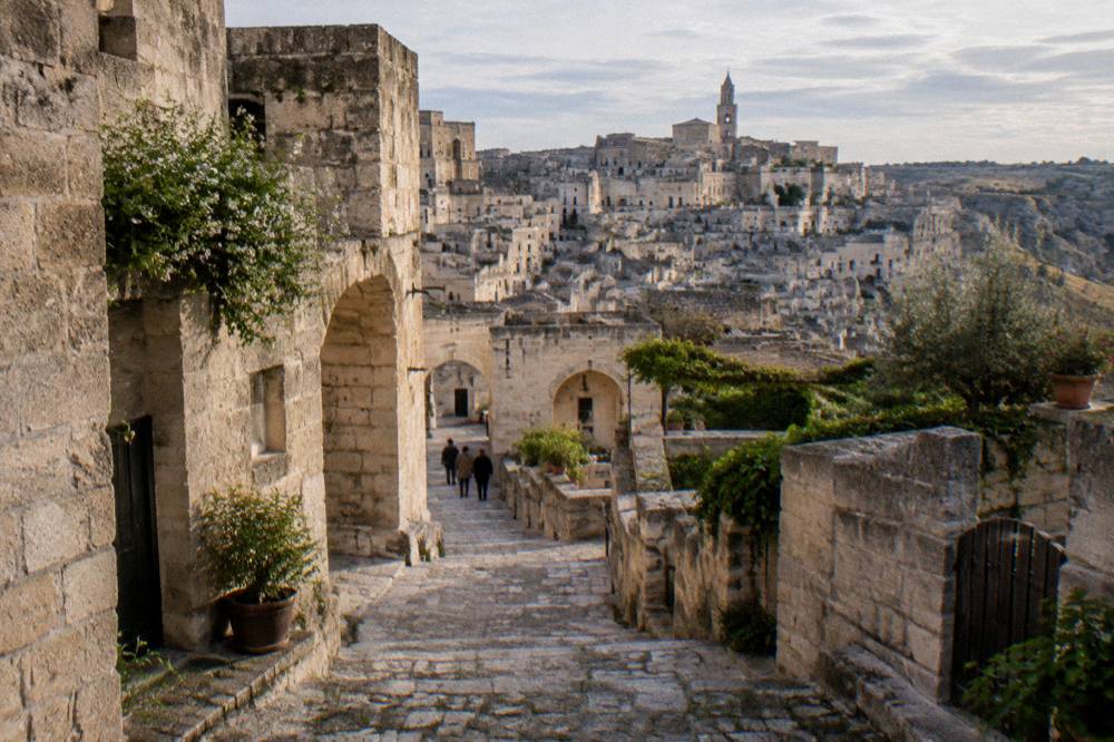 A stone lane stepping down between cave houses, arches and small gardens, a few tiny distant visitors, soft late light, Matera