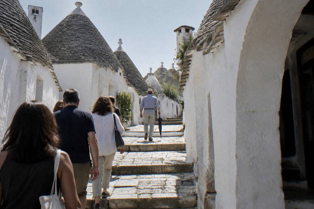 A group seen from behind walking up a stepped lane lined with white houses with gray conical roofs, a guide in front holding a closed umbrella, bright light, Alberobello