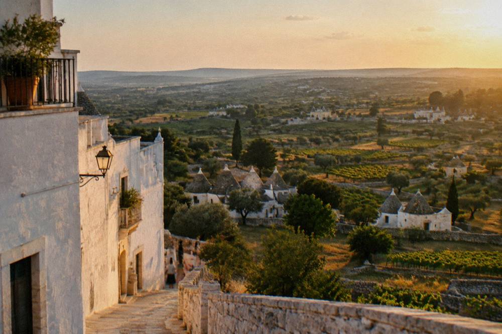 A valley of vineyards and dry-stone walls with scattered conical-roofed farm houses seen from the edge of a white hill town, hazy light, Locorotondo