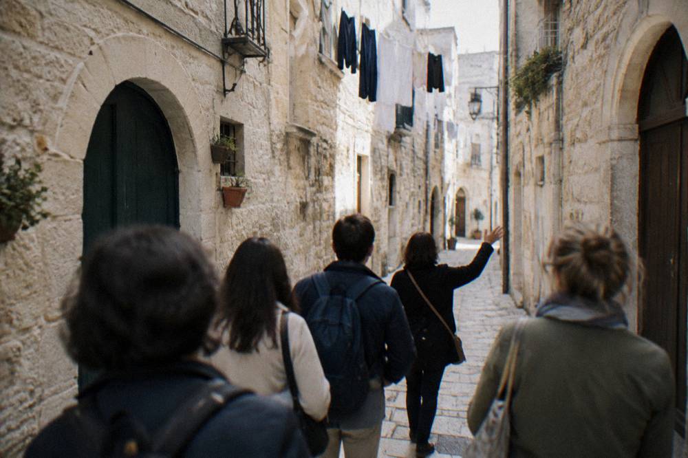 A small group seen from behind following a guide's raised hand along a pale limestone old-town lane, arched doorways and washing overhead, soft morning light, Bari Vecchia