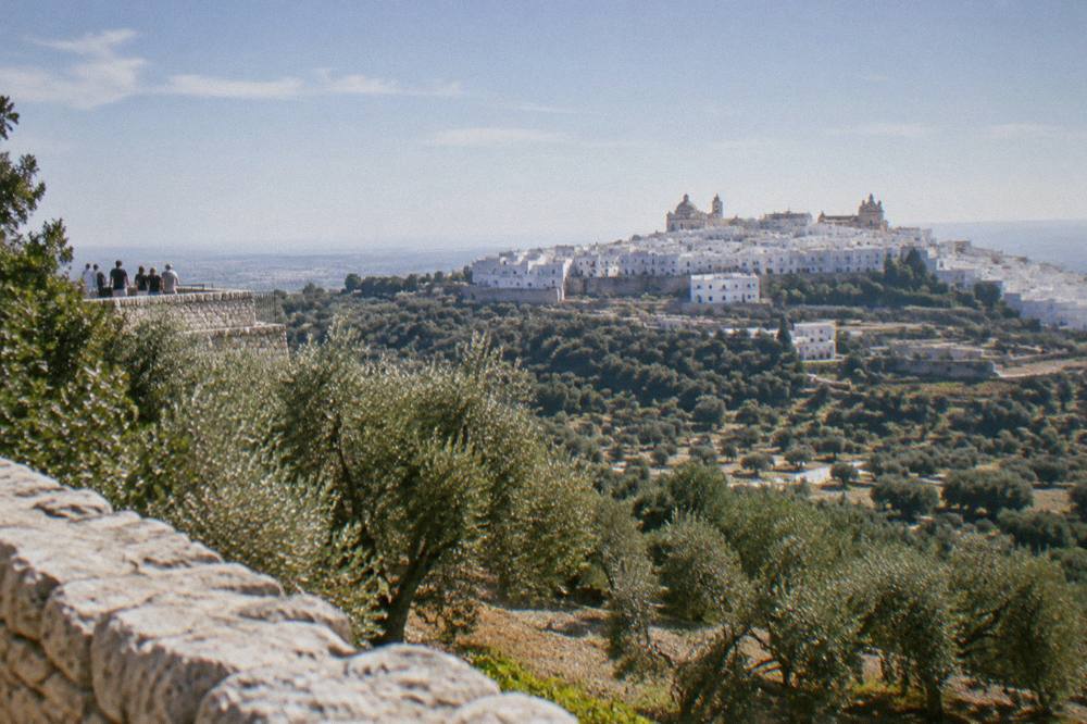 A dazzling white hill town on a ridge above silvery olive groves, a small group far off on a lookout, hazy sky, Ostuni