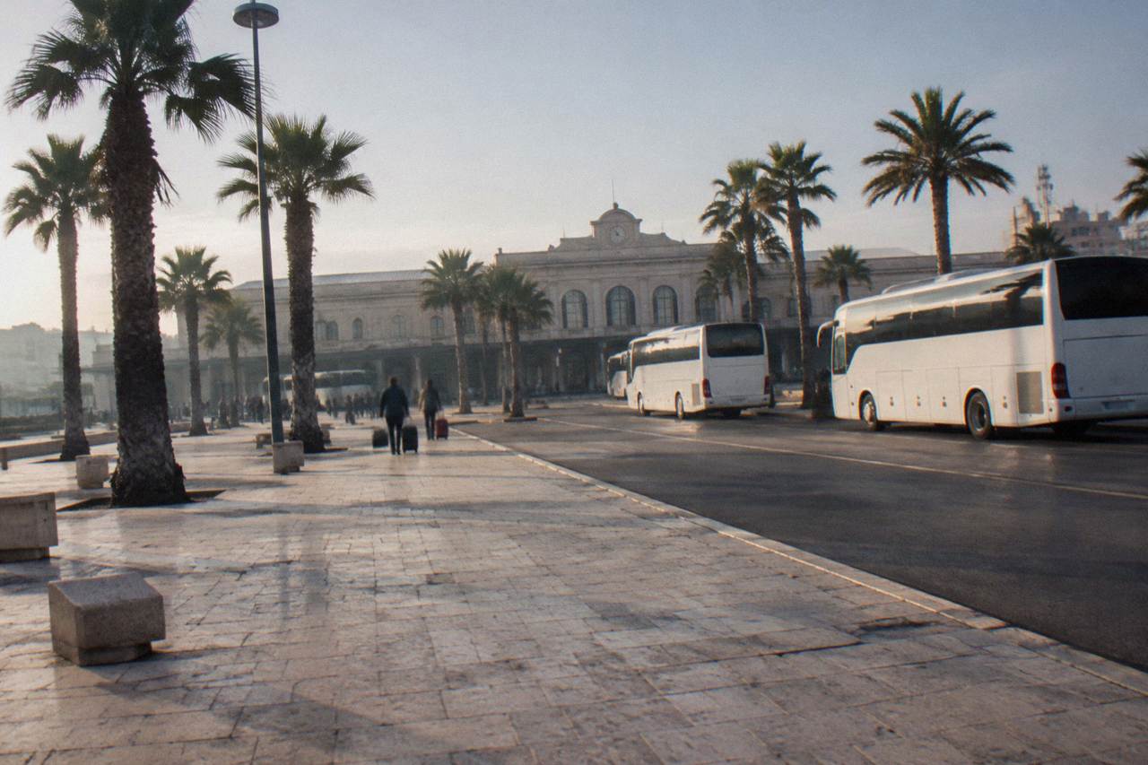 Station forecourt with plain white buses, palm trees and distant travellers with wheeled suitcases