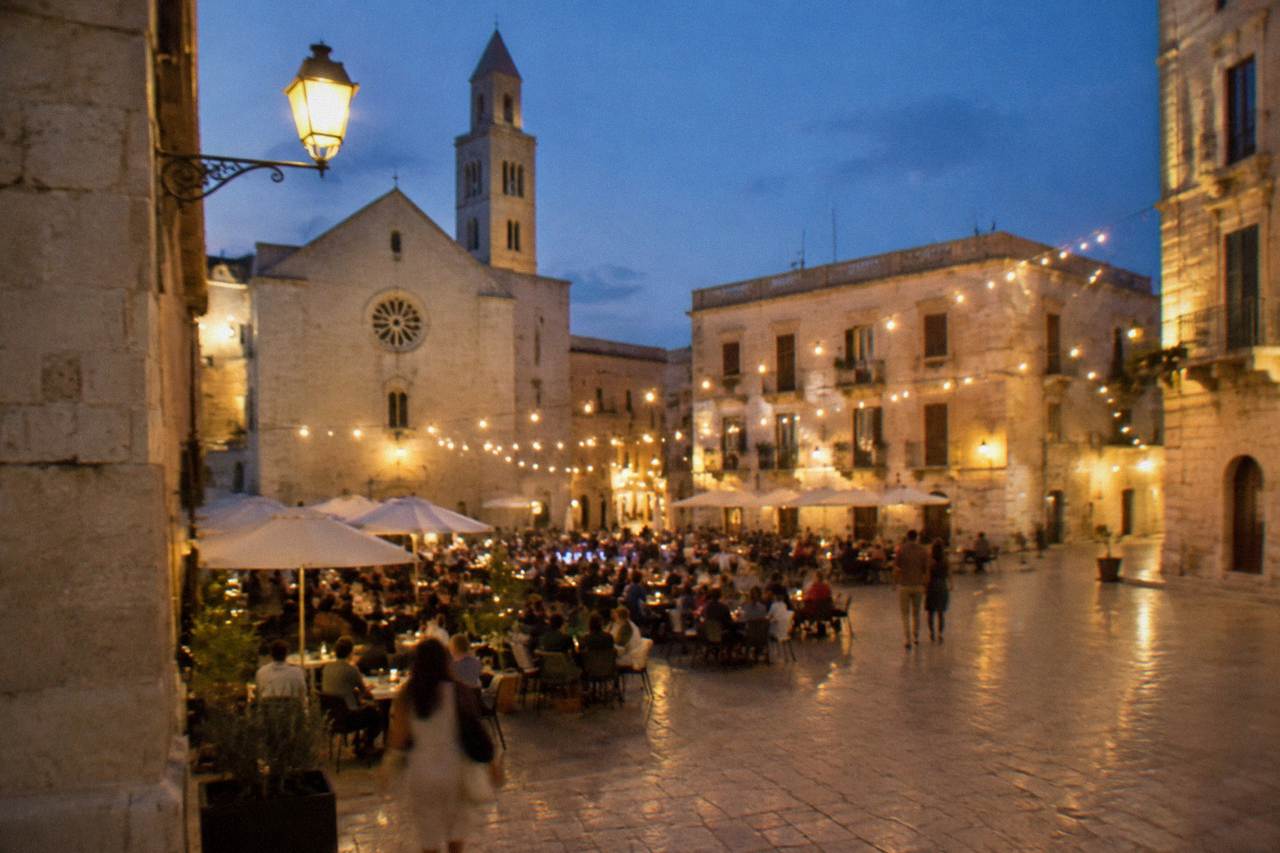 Pale limestone old town square at dusk with strings of lights and out-of-focus diners at cafe tables