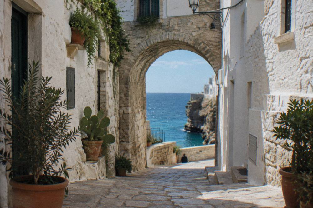 Narrow whitewashed cliff-top old town lane with a stone arch, potted plants and a glimpse of blue sea