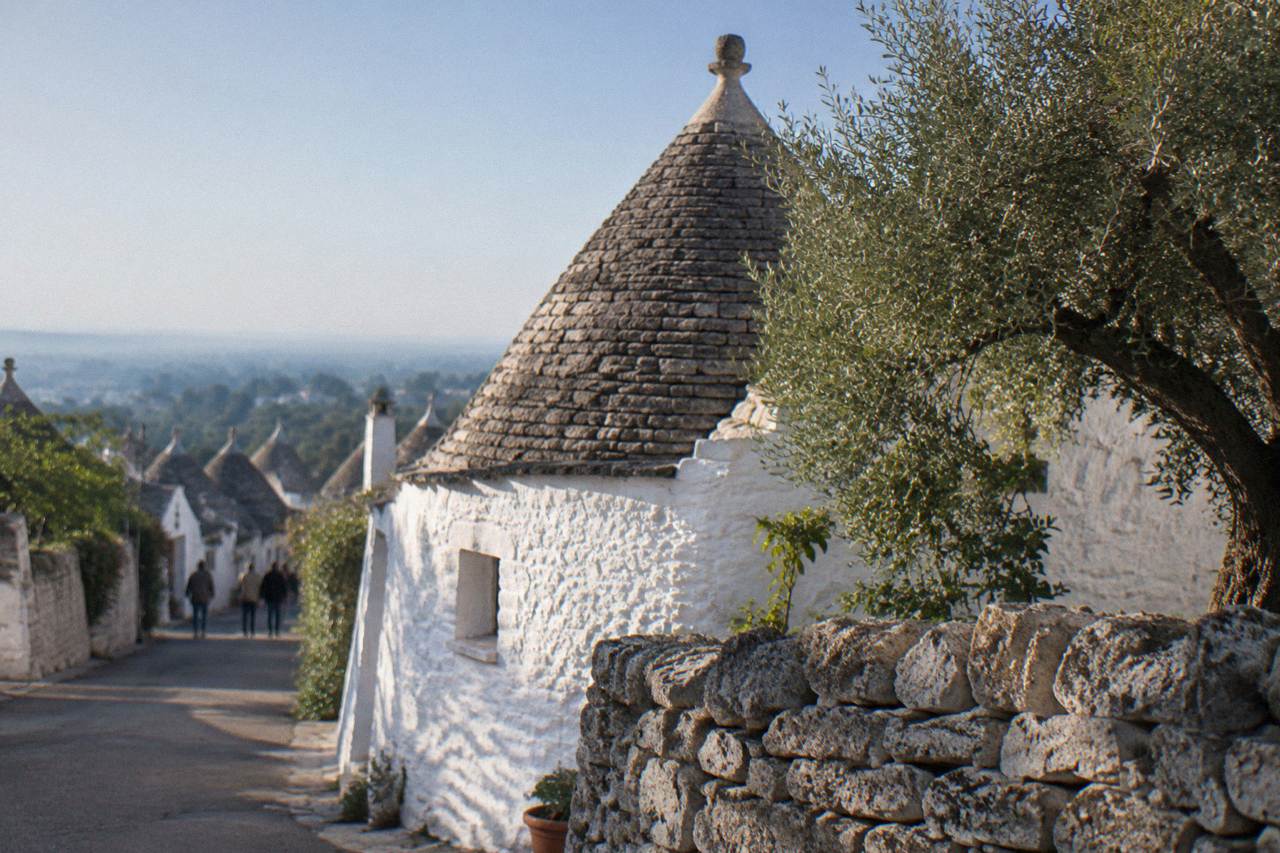 White house with a grey conical stone roof and a pinnacle behind a dry-stone wall and an olive tree