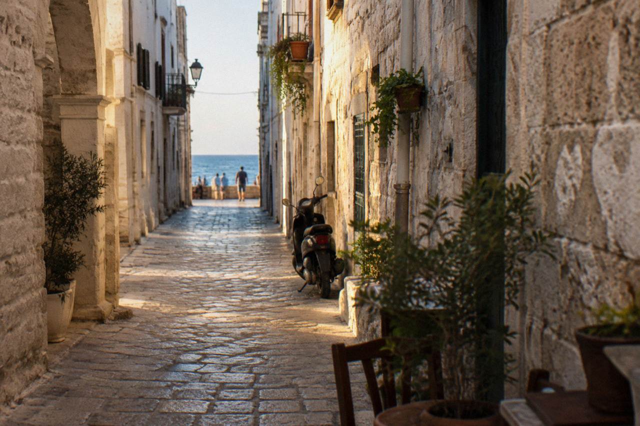 Straight old town lane between whitewashed walls ending in a bright slit of open sea with a parked scooter