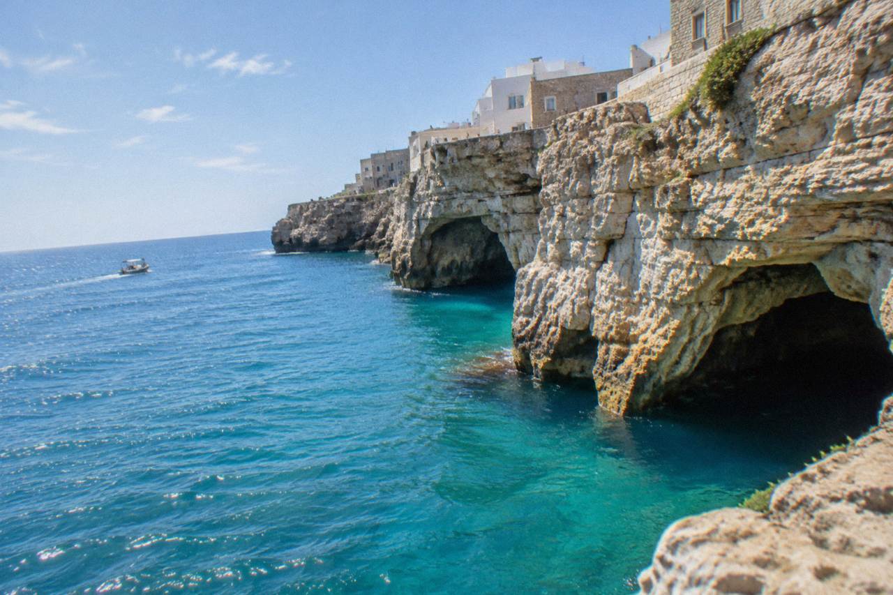 Sea cave openings at the base of tall limestone cliffs with turquoise water and a small boat far off