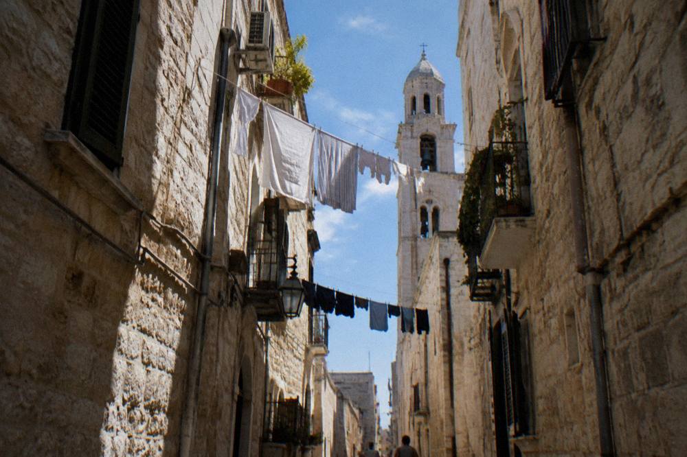 Looking up between close walls at a strip of blue sky crossed by washing lines and a bell tower