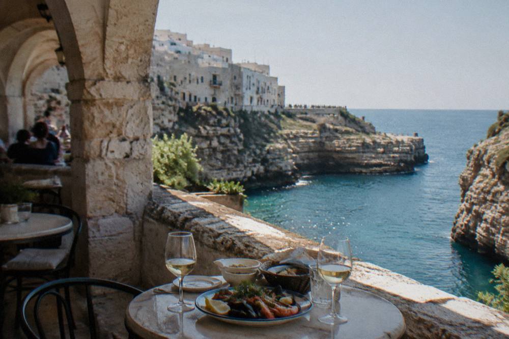 Small round tables on a cliff-edge terrace with glasses and a plate of seafood above blue water