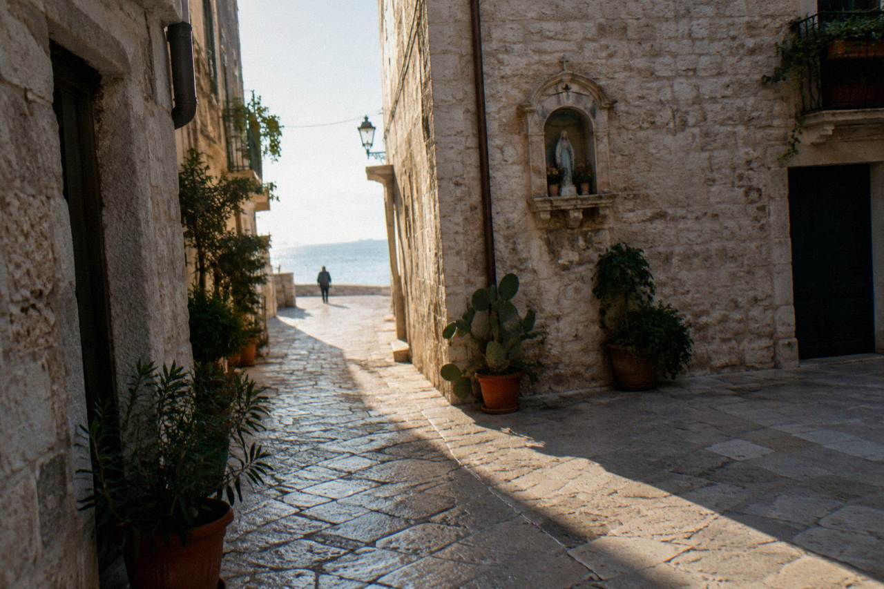 Crossing of narrow old town lanes with a stone shrine niche, potted plants and sunlight slicing between buildings