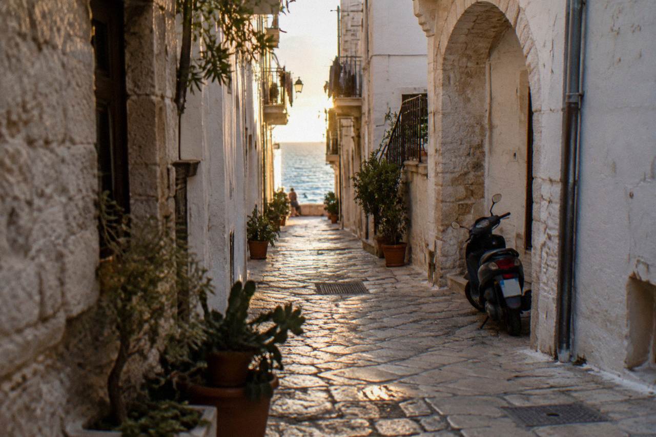 Straight old town lane between whitewashed walls ending in a bright slit of open sea with a parked scooter