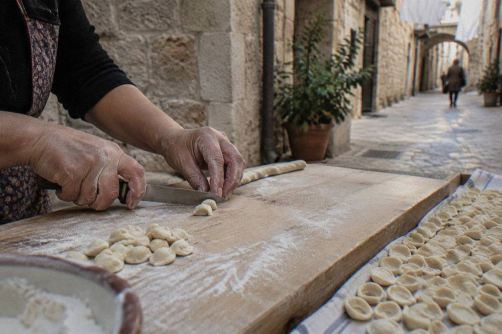 Floury hands drawing a small piece of dough across a board with a knife beside rows of finished pasta