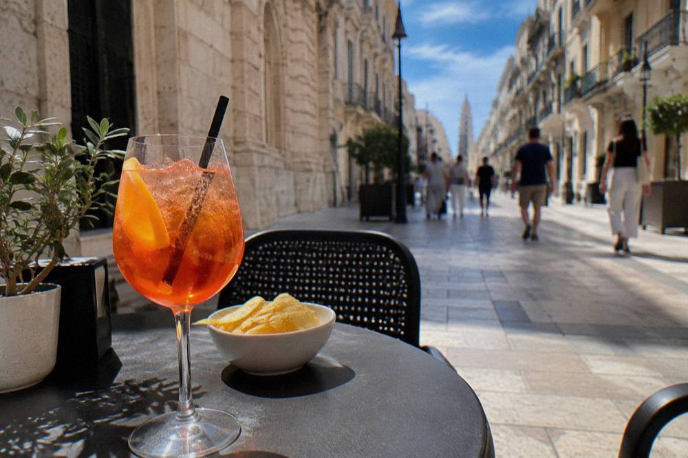 Pavement cafe table on a pedestrian street with an orange spritz and a small plate of crisps, passers-by blurred