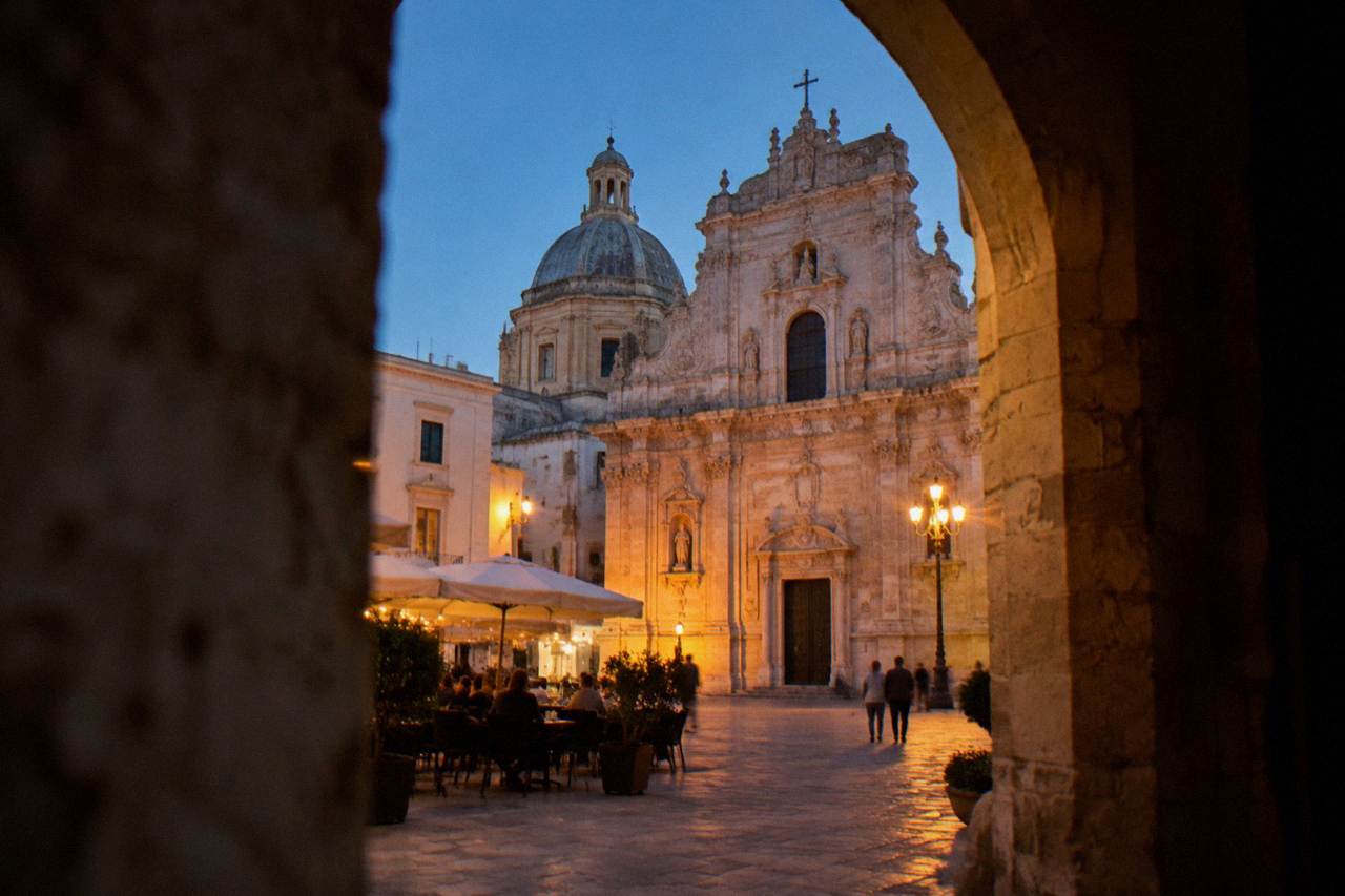 Baroque cathedral facade in warm pale stone on a small square with a dome behind and cafe tables